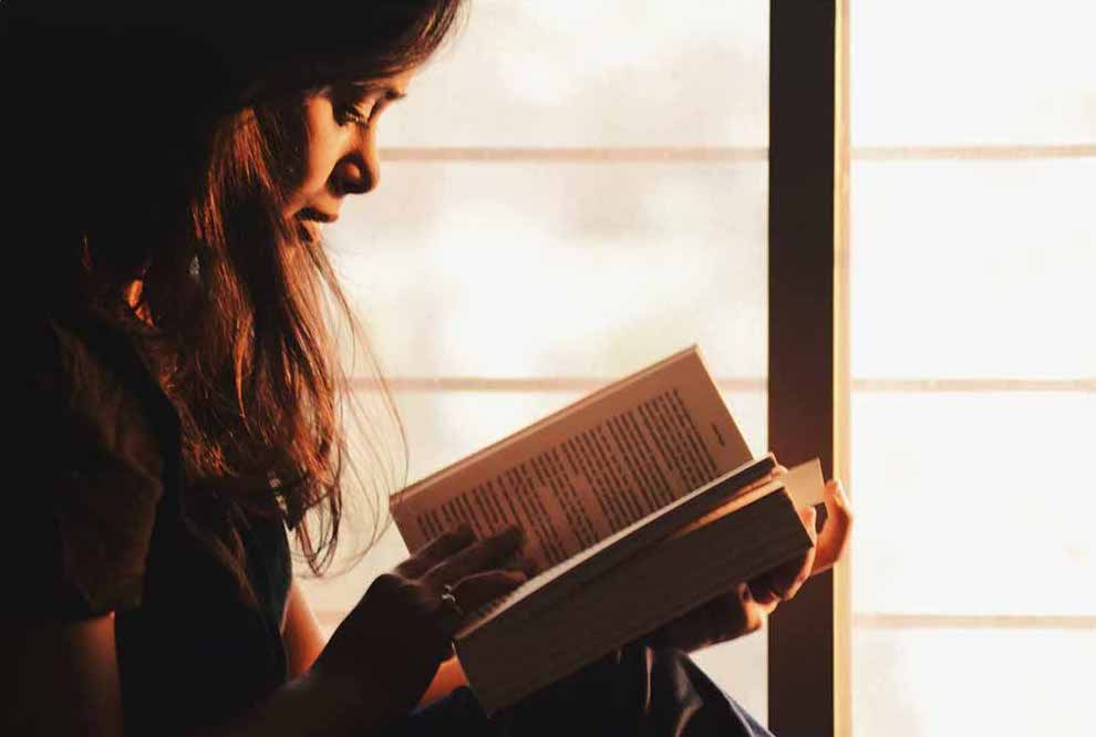 A girl reading a book near the windows.